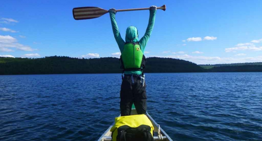 a person stands at the front a canoe and raises their paddle in the air on an outward bound course